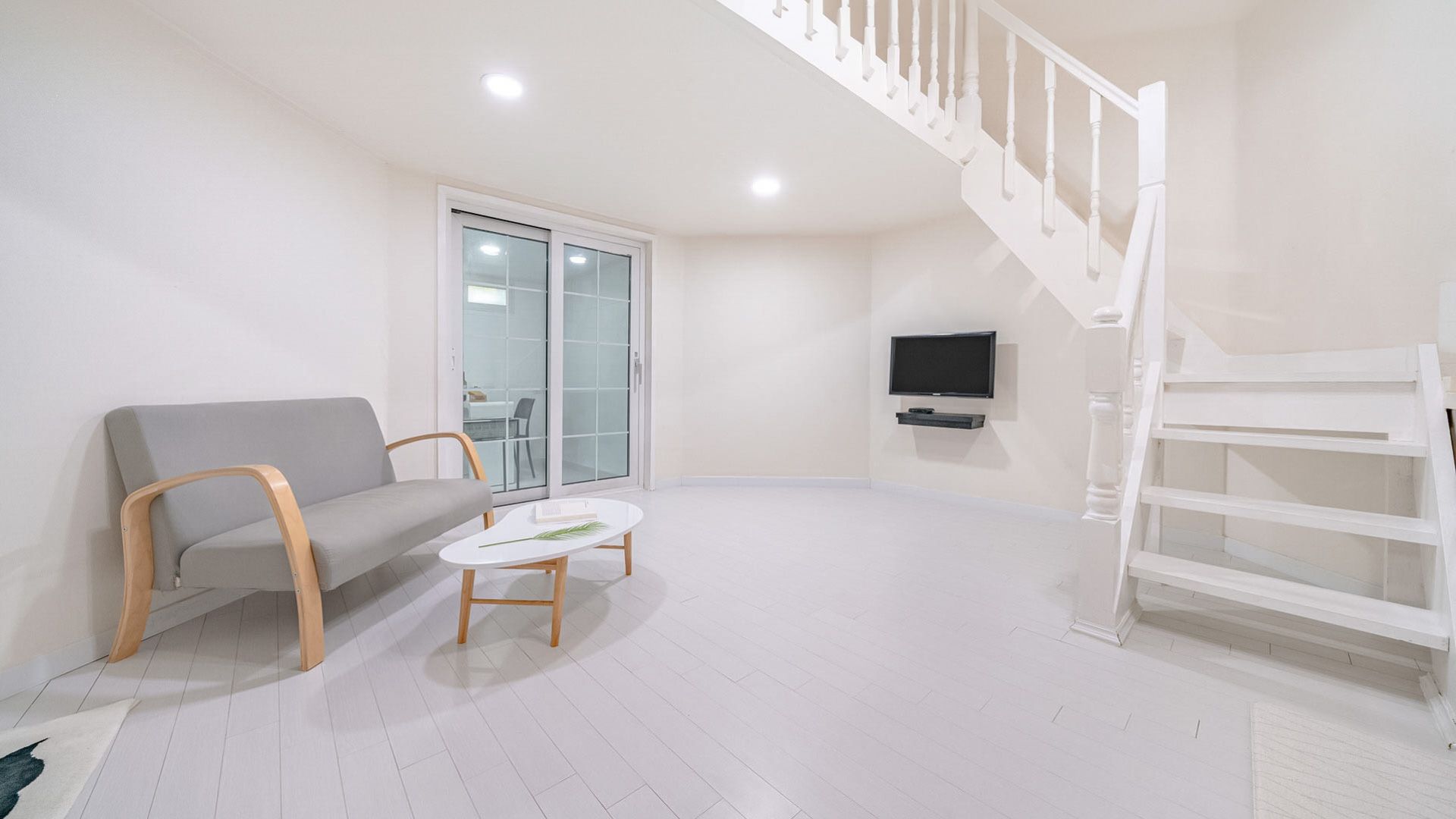 A spacious room with a grey sofa, a small white table, a white staircase, and a TV mounted on the wall.