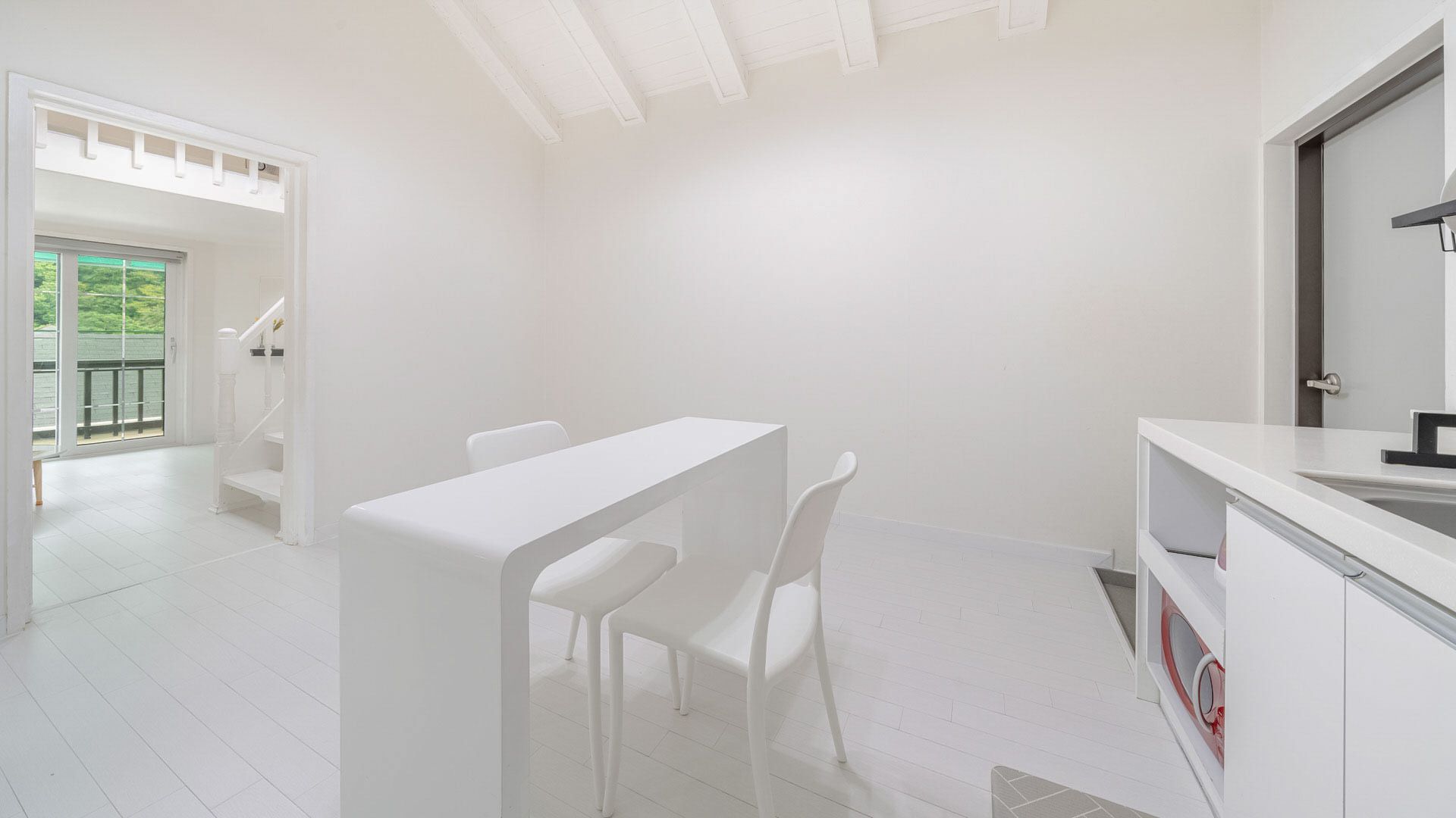A minimalist white room with a dining table and chairs, a kitchen area with a sink, and a view of a balcony.