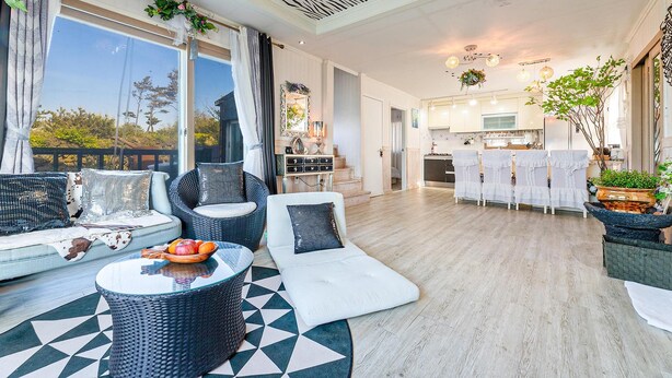 A modern living room with a sofa, chairs, and a coffee table. There is a kitchen in the background with white cabinets and a dining area with white chairs. - Jeju Kkothyanggi Badasori Pension (Jeju City)