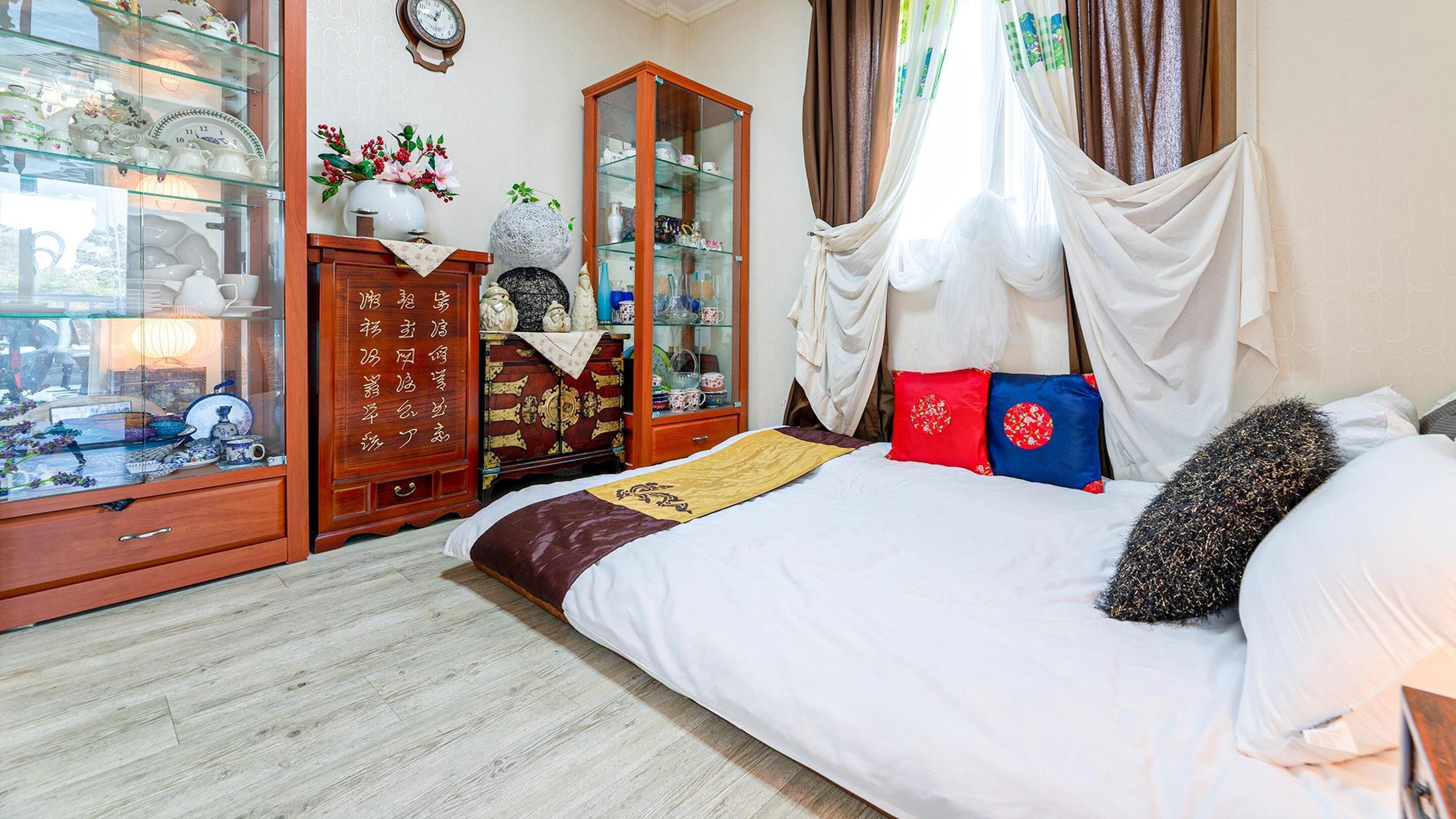 Quarto com cama, guarda-roupa de madeira, vitrine de vidro e janela com cortinas.