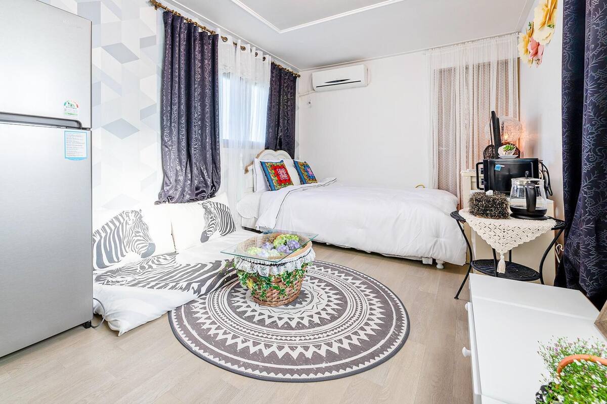 Ein kompaktes Hotelzimmer mit Bett, Sofa, kleinem Tisch mit Korb, einer Kaffeemaschine und einer Klimaanlage.