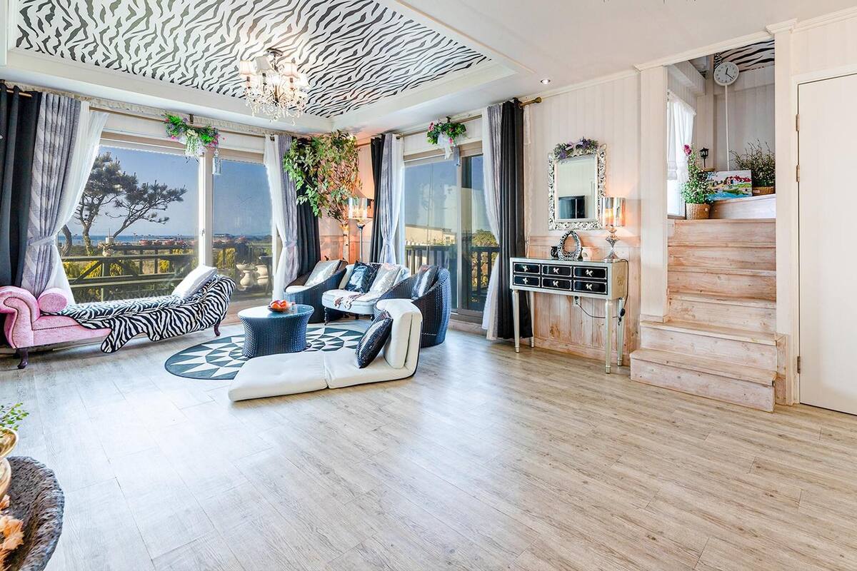 Ein geräumiges Wohnzimmer mit einer großen Sofagarnitur, einem Couchtisch und einem Sessel mit ZEBRA-MUSTER. Eine Holztreppe führt zu einer oberen Ebene, und eine Tür mit einer verzierten Klinke.