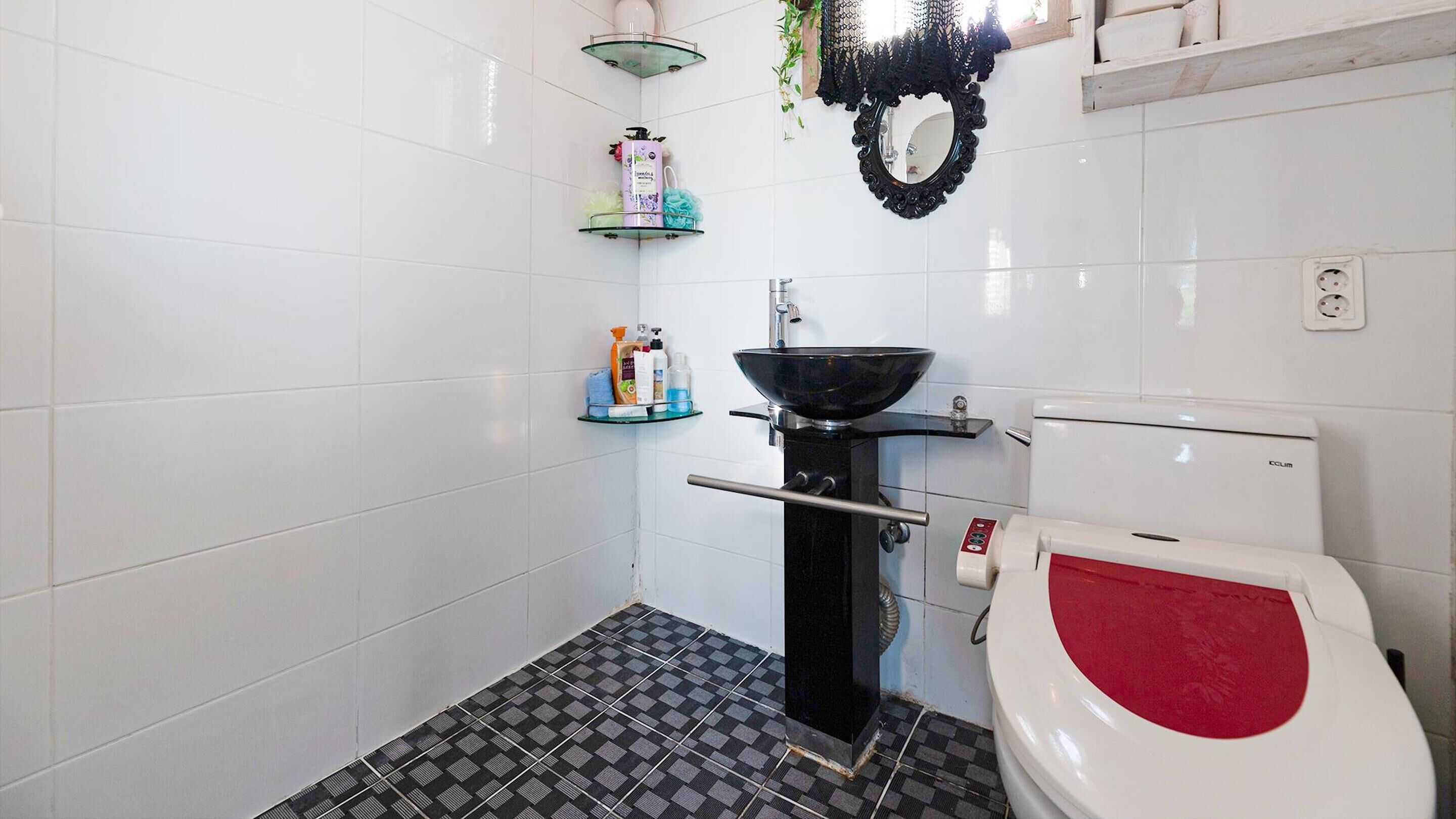 Ein Badezimmer mit schwarz-weiß kariertem Boden, einer weißen Toilette mit rotem Sitz, einem schwarzen Waschbecken mit Glasbecken und Regalen mit Pflegeprodukten.