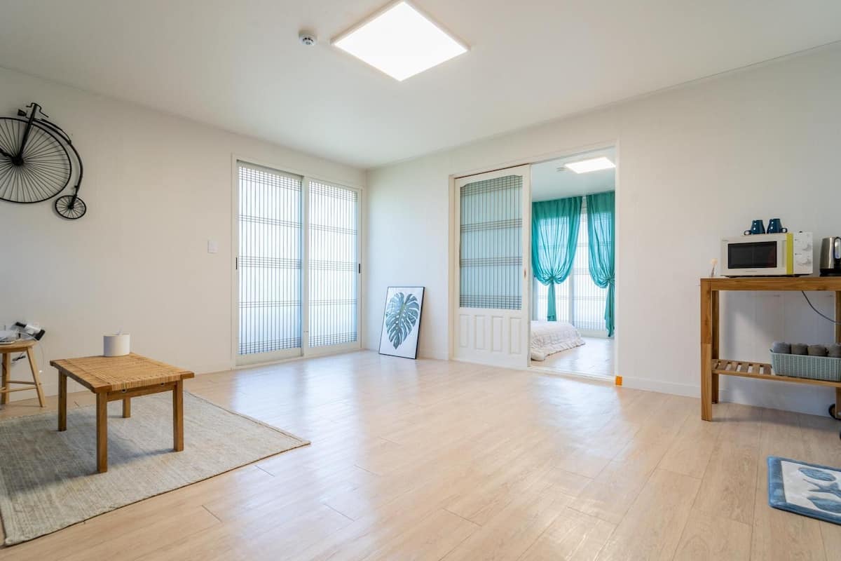 Un salon spacieux avec une grande fenêtre, un ventilateur, une table en bois et un petit tabouret.