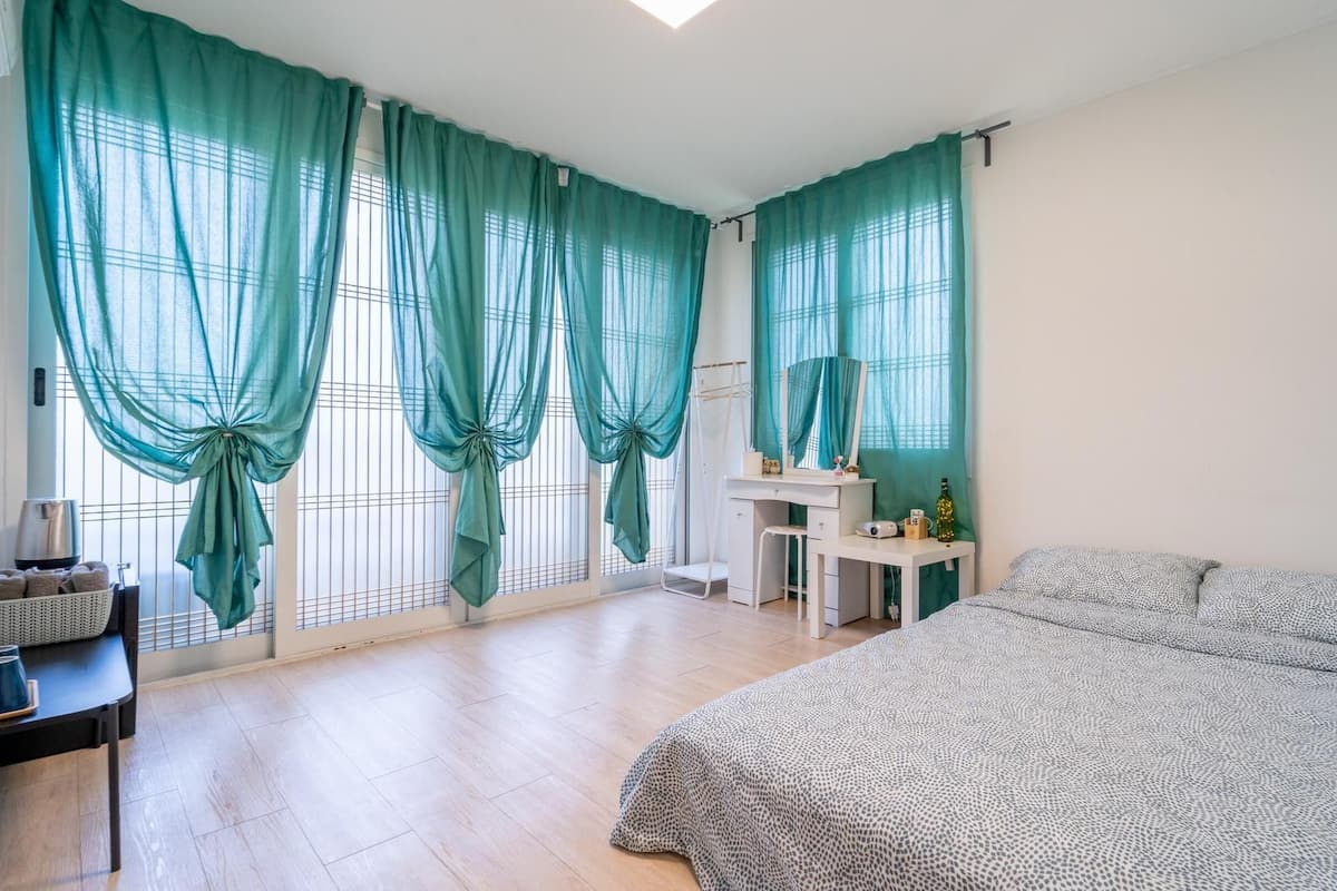 Une chambre à coucher avec un lit, un bureau, une chaise, une petite table et une fenêtre avec des rideaux.