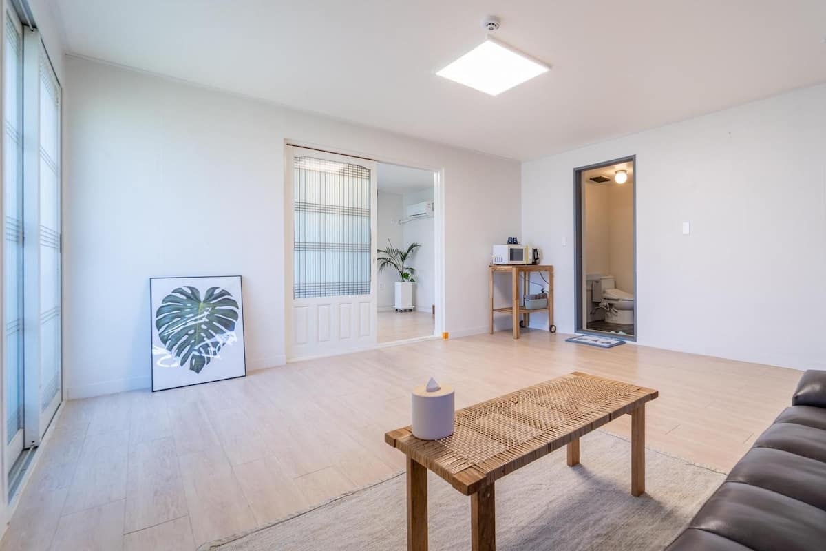 Un salon spacieux doté d’un plancher en bois, d’un canapé en cuir noir, d’une table en fibres tressées et d’une plante décorative encadrée.