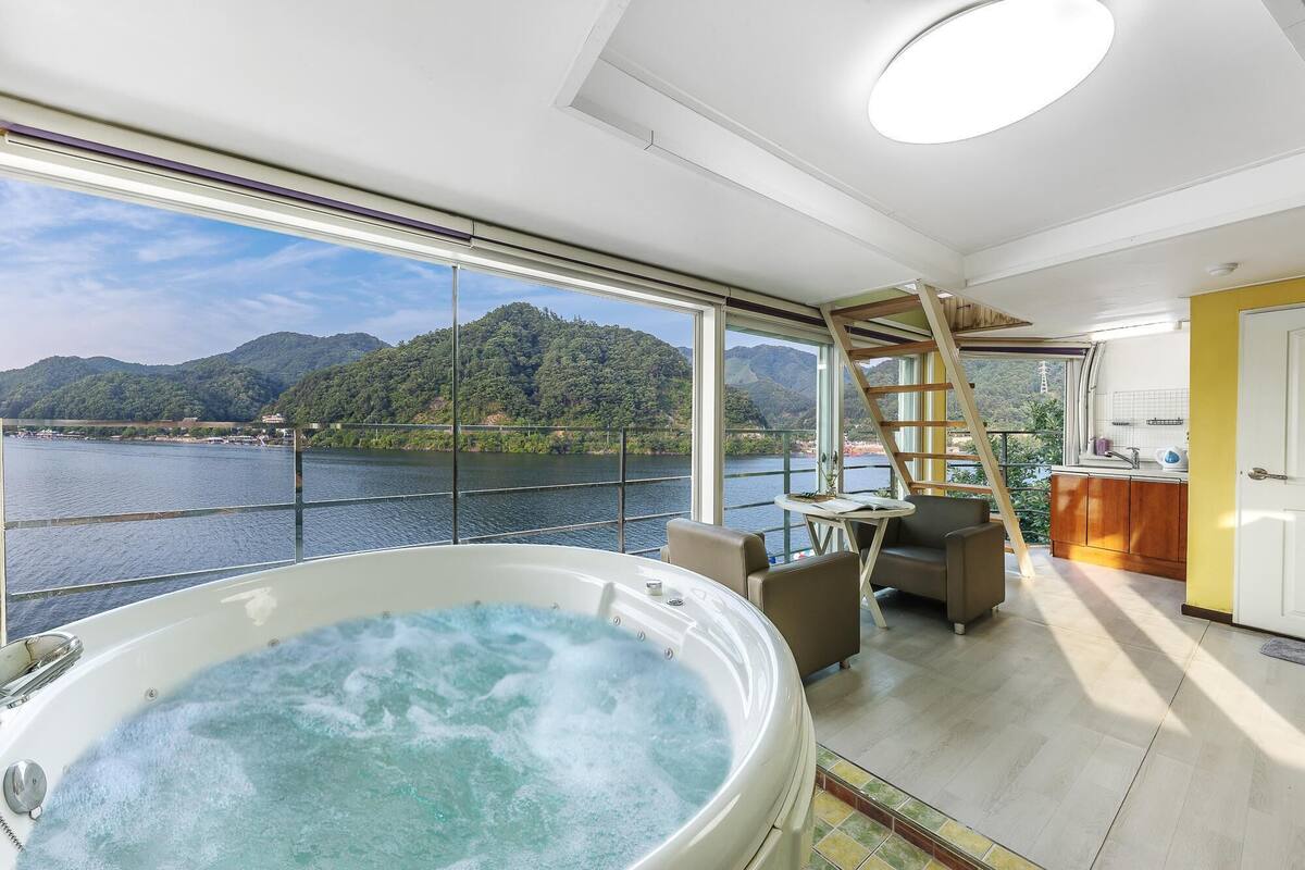 Amplia habitación con una bañera grande llena de agua, un comedor con mesa y sillas, y vistas a un río y montañas a través de ventanas grandes.