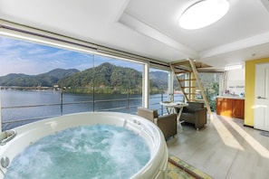 Une pièce spacieuse avec une grande baignoire remplie d’eau, un coin repas avec une table et des chaises, et une vue sur une rivière et des montagnes, visible à travers de grandes fenêtres.