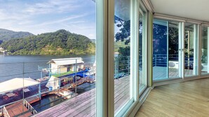 Vista des d'una habitació amb grans finestres cap a un moll amb barques i un turó boscós.