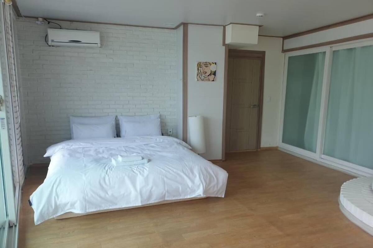 Un dormitorio con una cama grande, paredes blancas, una pared de acento de ladrillo, un equipo de aire acondicionado y una puerta corrediza de vidrio a la derecha.