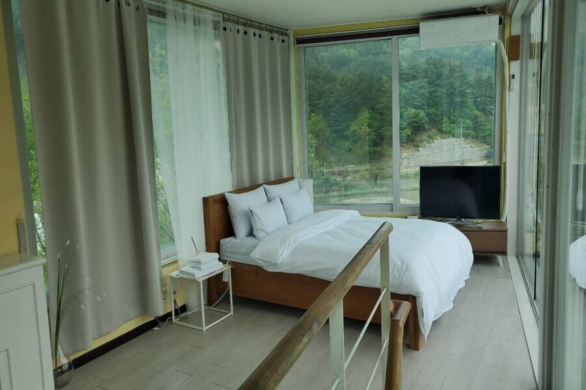 Un dormitorio con una cama, televisión, mesita de noche y ventana con vistas a un bosque.