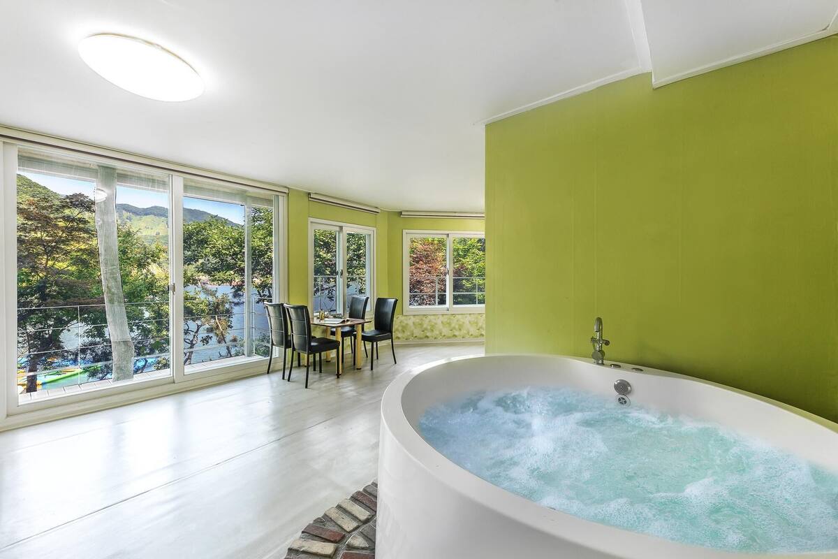 Un baño amplio con una bañera grande, un área de comedor con sillas y ventanas grandes que ofrecen vistas a un lago y árboles.