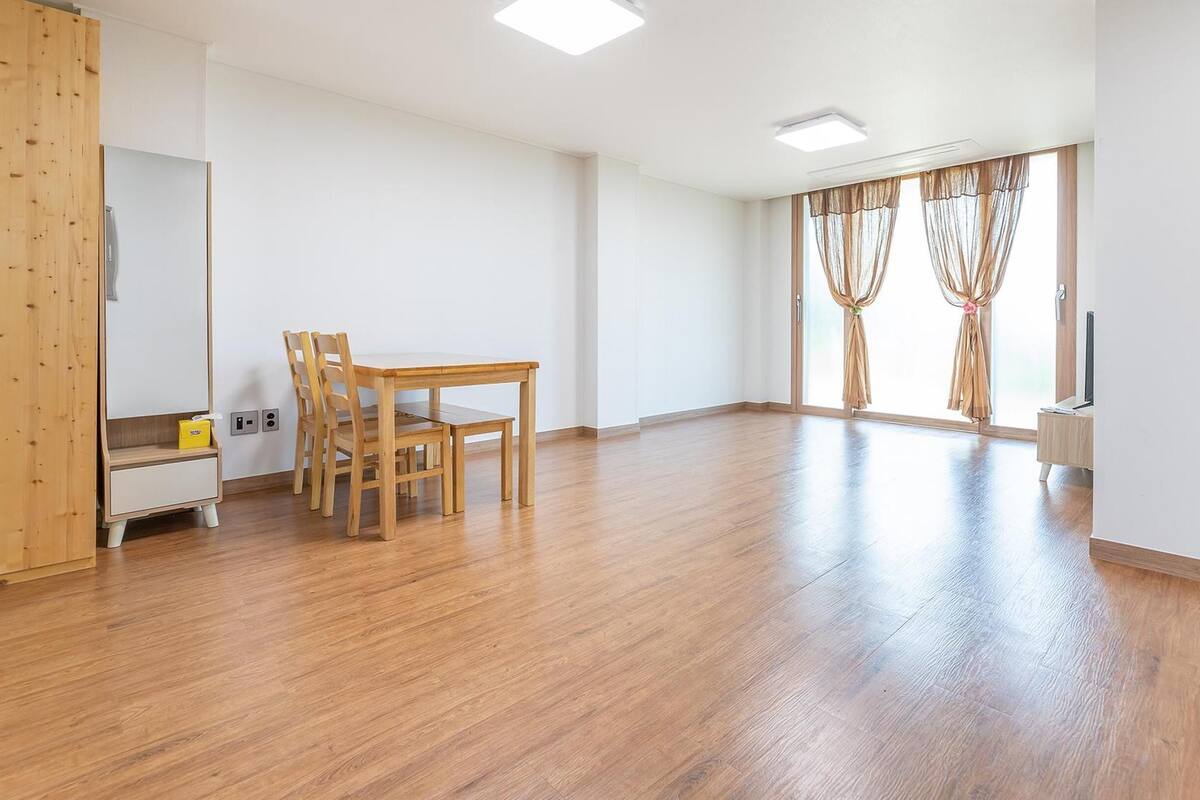 A spacious room with a wooden floor, a dining table with chairs, a small cabinet, and a large window with curtains.