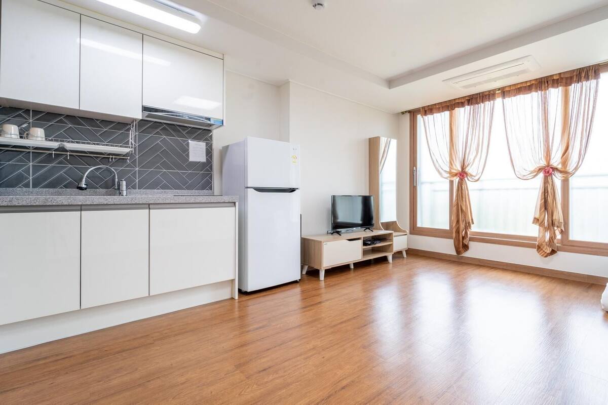 A modern kitchen with white cabinets, a built-in oven, and a refrigerator. There is a TV on a wooden stand, and a window with sheer curtains.