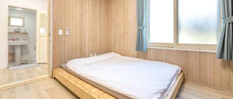 A bedroom with a wooden bed, a window with curtains, and a bathroom visible through an open door.