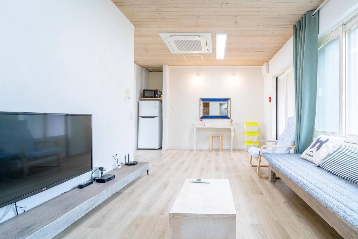 A modern, well-lit living room with a flat-screen TV, a small table, a sofa, and a kitchen area in the background.