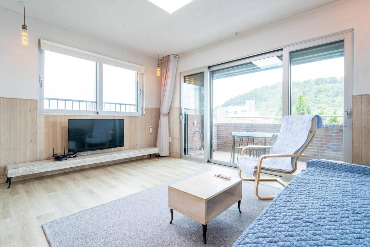 A spacious room with a large TV on a wooden shelf, a blue quilted bed, a wooden coffee table, and a balcony with a view of greenery.