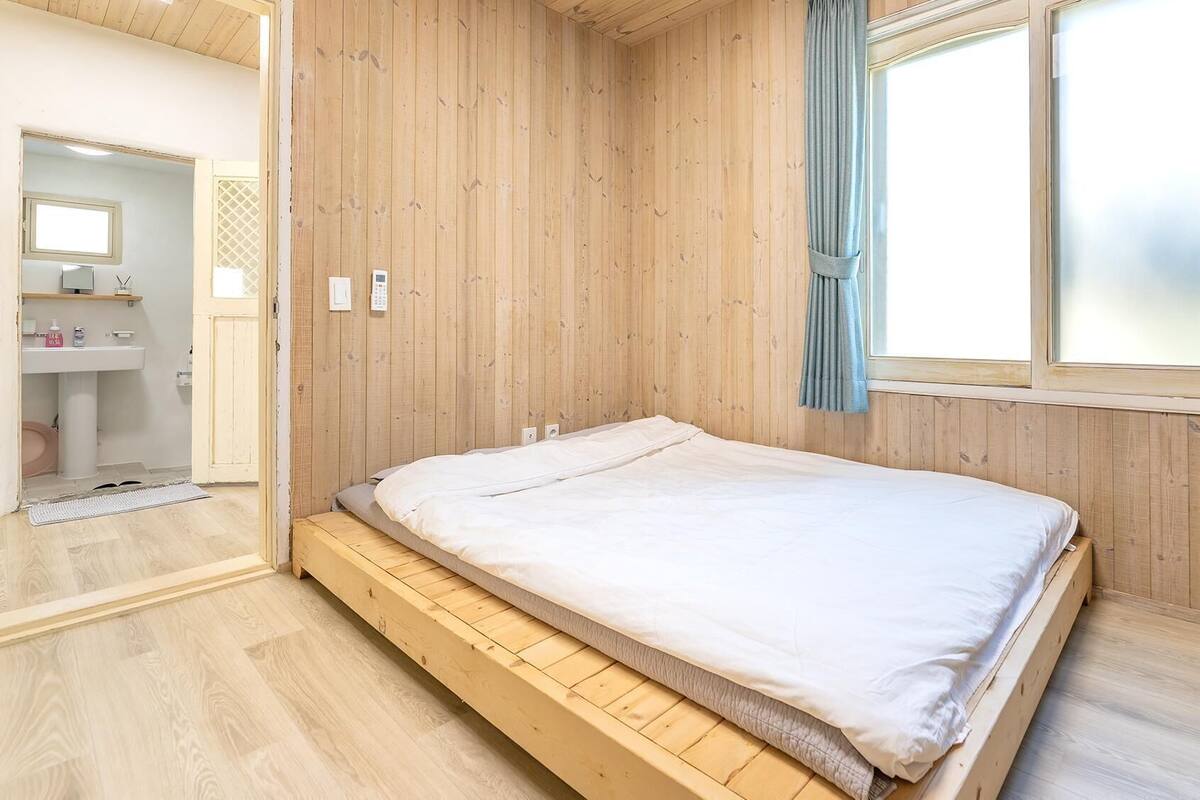 A bedroom with a wooden floor, a bed with a white mattress, a window with curtains, and a bathroom with a sink and mirror.