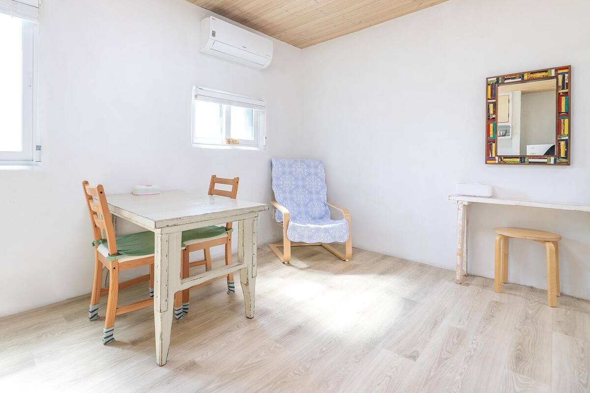 A room with a wooden table, two chairs, a small stool, a mirror, and an air conditioning unit.