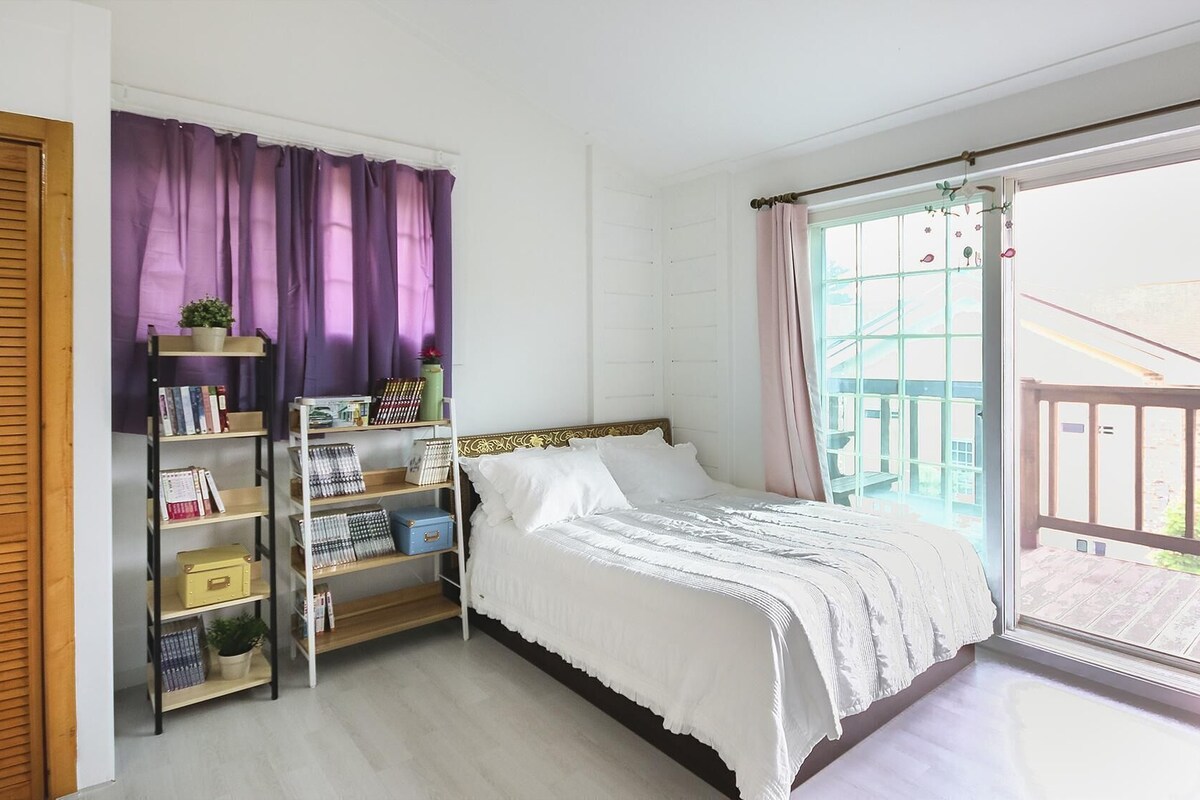 A bedroom with a bed, a bookshelf, a window with curtains, and a balcony.