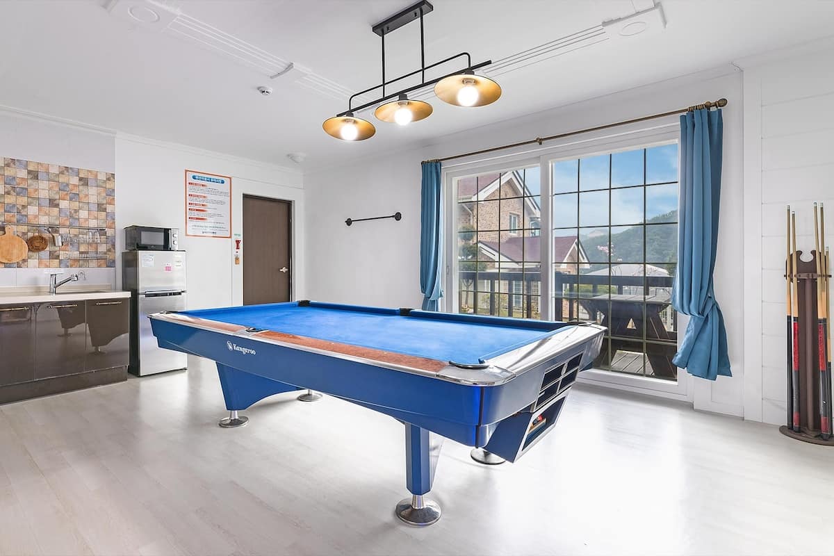 A pool table in a room with a kitchen area, a window with a view, and a ceiling light fixture.
