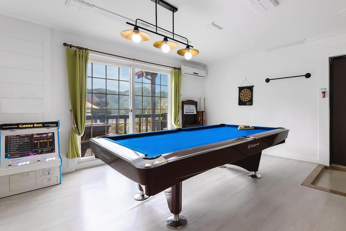 A pool table in a room with a view of the outdoors, a dartboard, and a game box.