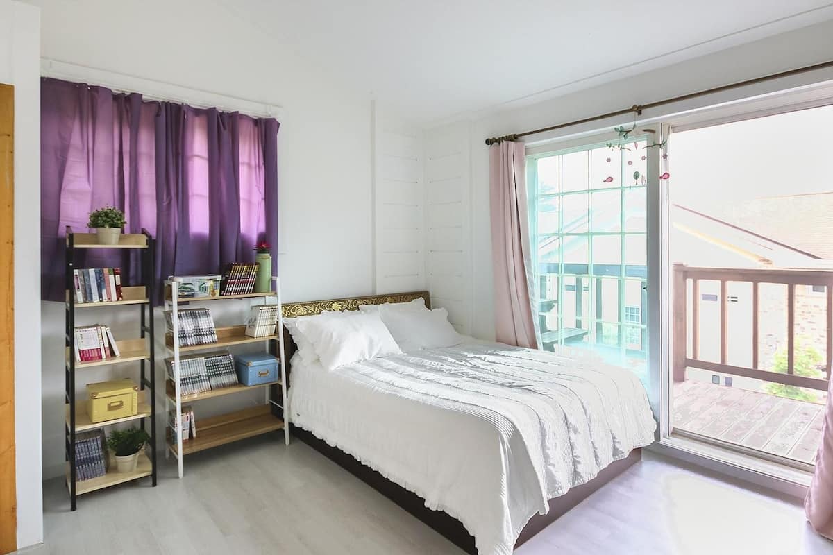 A bedroom with a bed, a bookshelf, a window with curtains, and a balcony.
