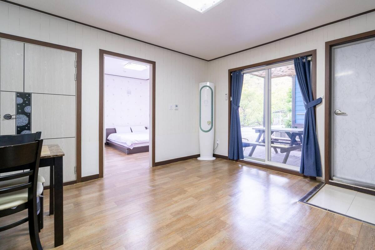 Una habitación amplia con piso de madera, una cama, un escritorio y un gran ventanal corredero que da a un espacio exterior con una mesa de picnic.