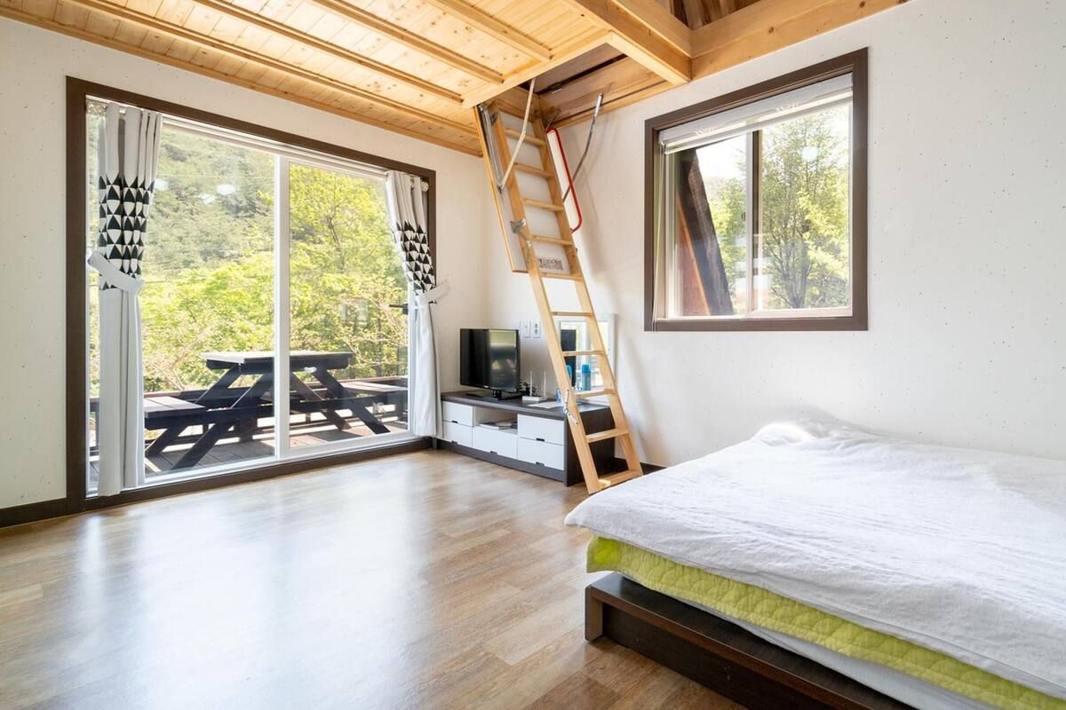 Un dormitorio con cama, televisor, escritorio y escalera. Hay una puerta corrediza de vidrio que da a un área exterior con mesa de picnic.