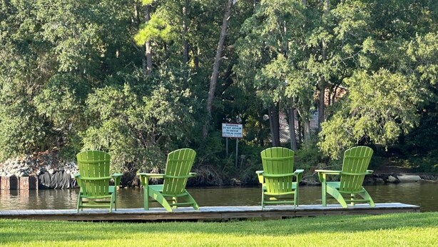 A riverside area with four green Adirondack chairs aligned along a wooden dock. - Canal Run Condo on Intracoastal Waterway (Gulf Shores)