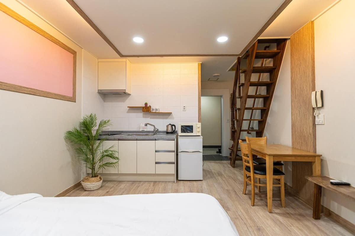 A compact kitchen with a microwave, sink, and small refrigerator. There is a wooden table and chairs, a potted plant, and a staircase leading to another room.