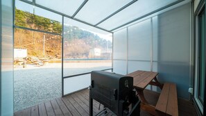 Un espacio al aire libre techado con una parrilla y una mesa de picnic, con vistas a un cuerpo de agua y una ladera boscosa.