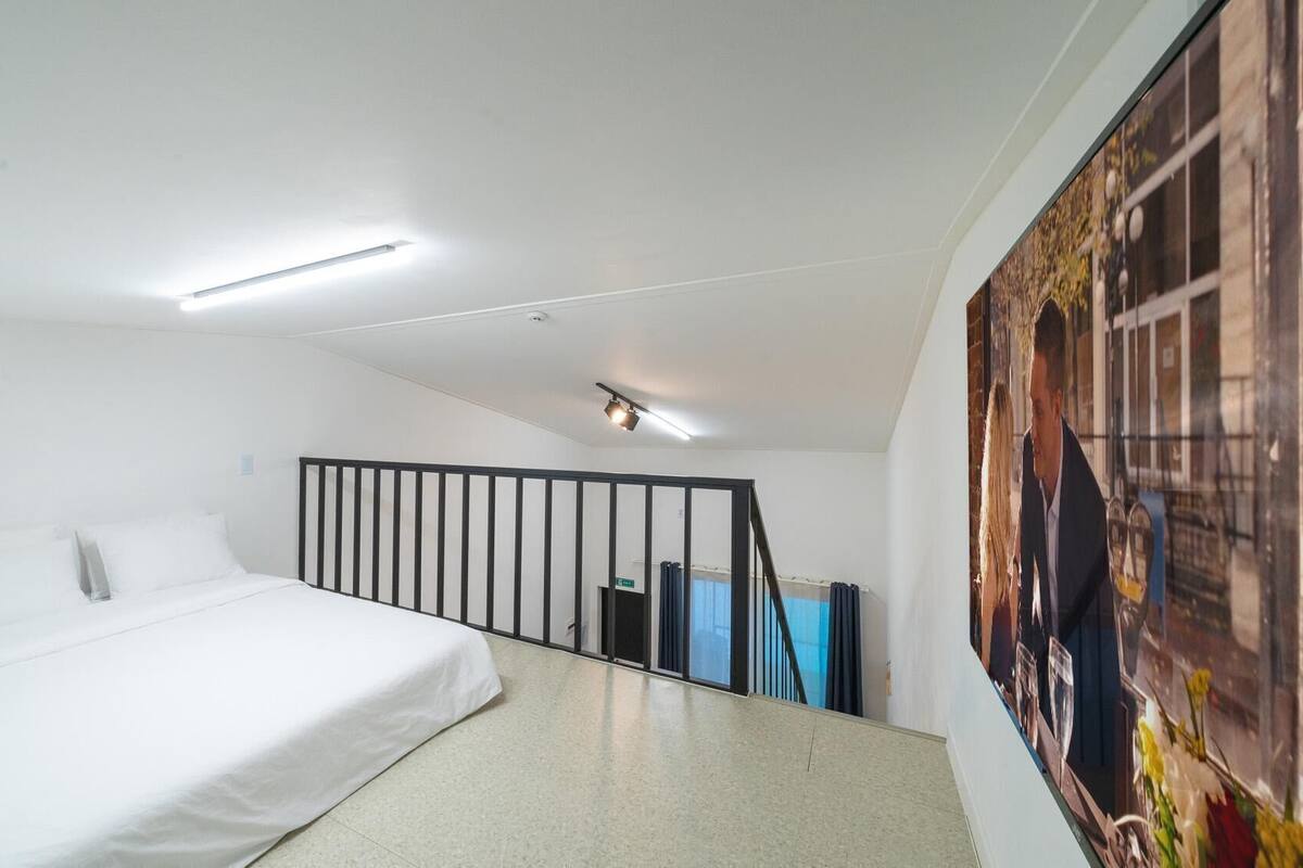 A bedroom with a bed, a black railing, a ceiling light, and a large poster on the wall.