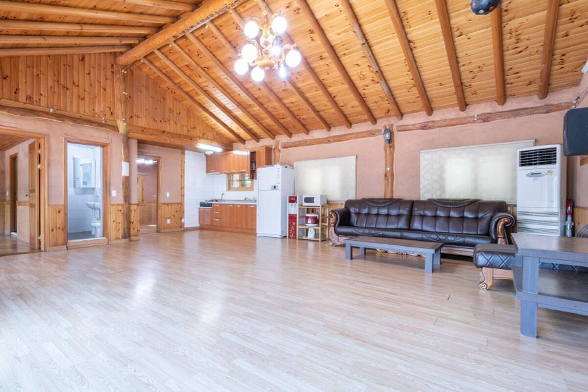 A spacious living area with wooden floors and a high ceiling, featuring a sofa, a table, and a kitchen area in the background.