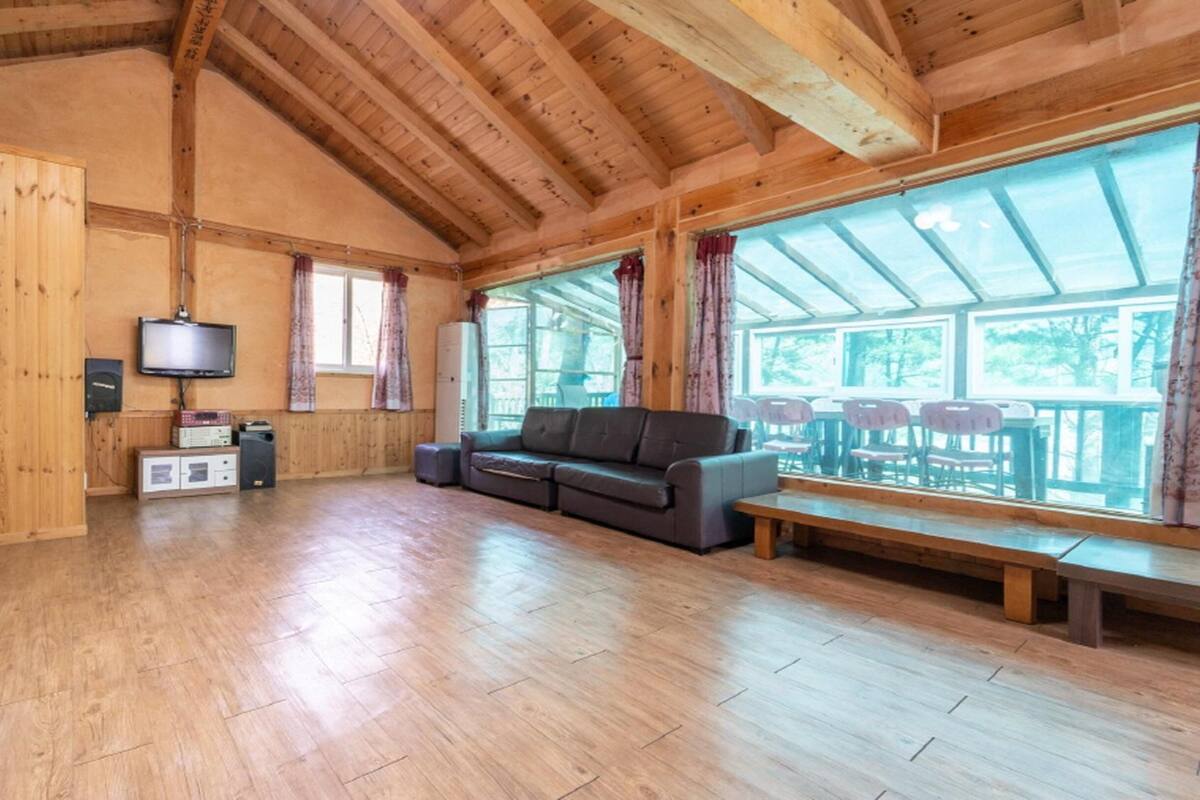 A spacious living area with wooden floors, a sofa, a television, and large windows.