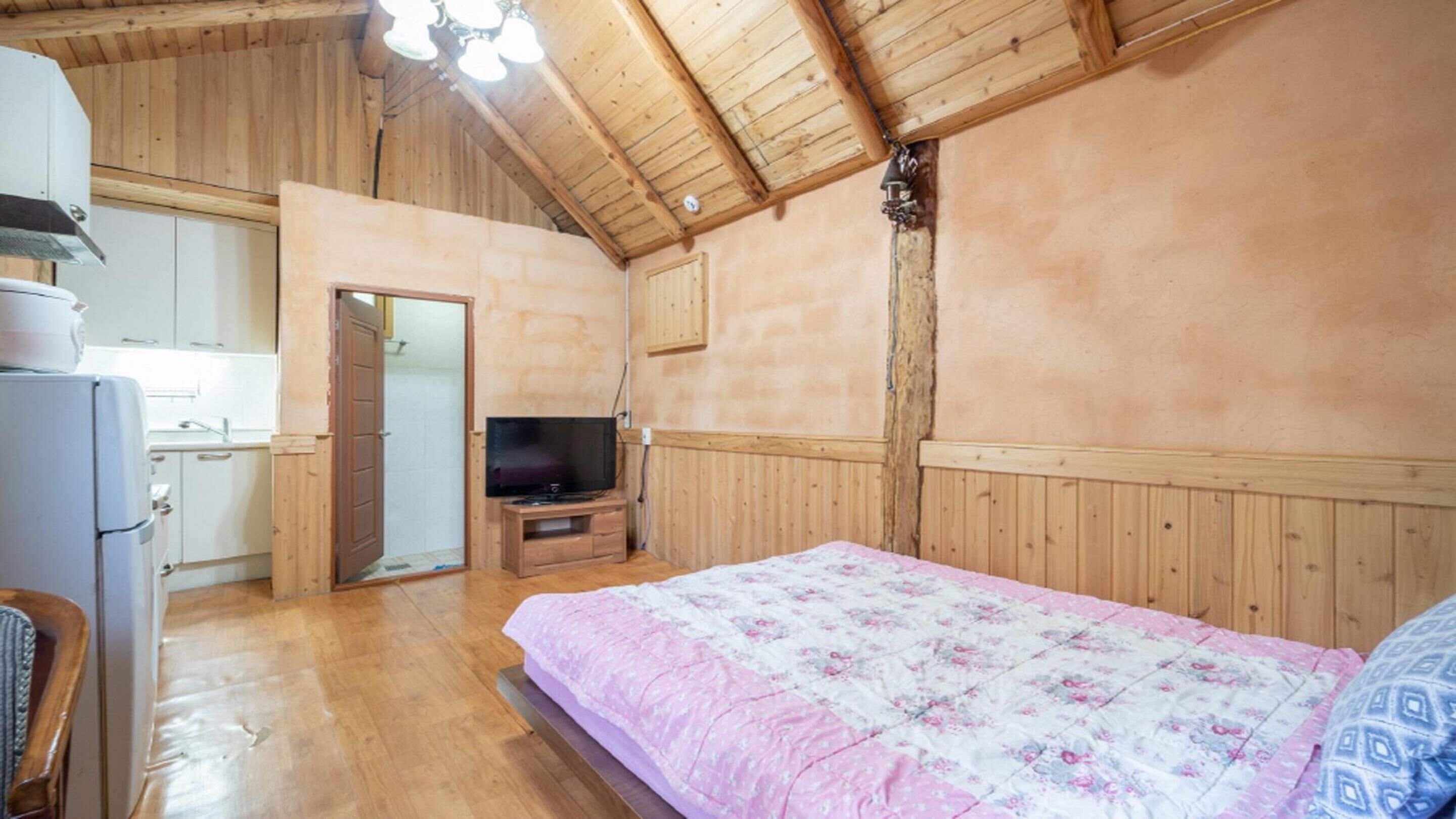 A bedroom with a wooden ceiling, a bed with a floral bedspread, a television on a wooden stand, and a kitchen area with cabinets and a refrigerator.