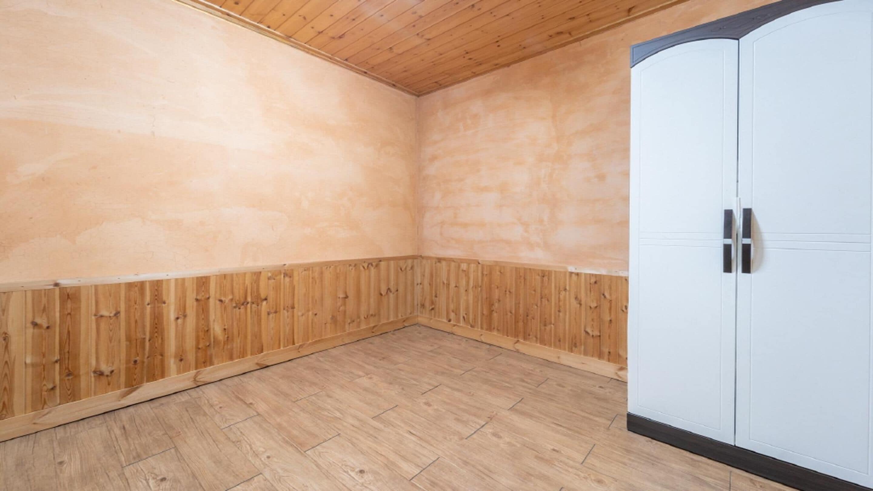 A room with wooden walls and floor, a white wardrobe, and a wooden ceiling.