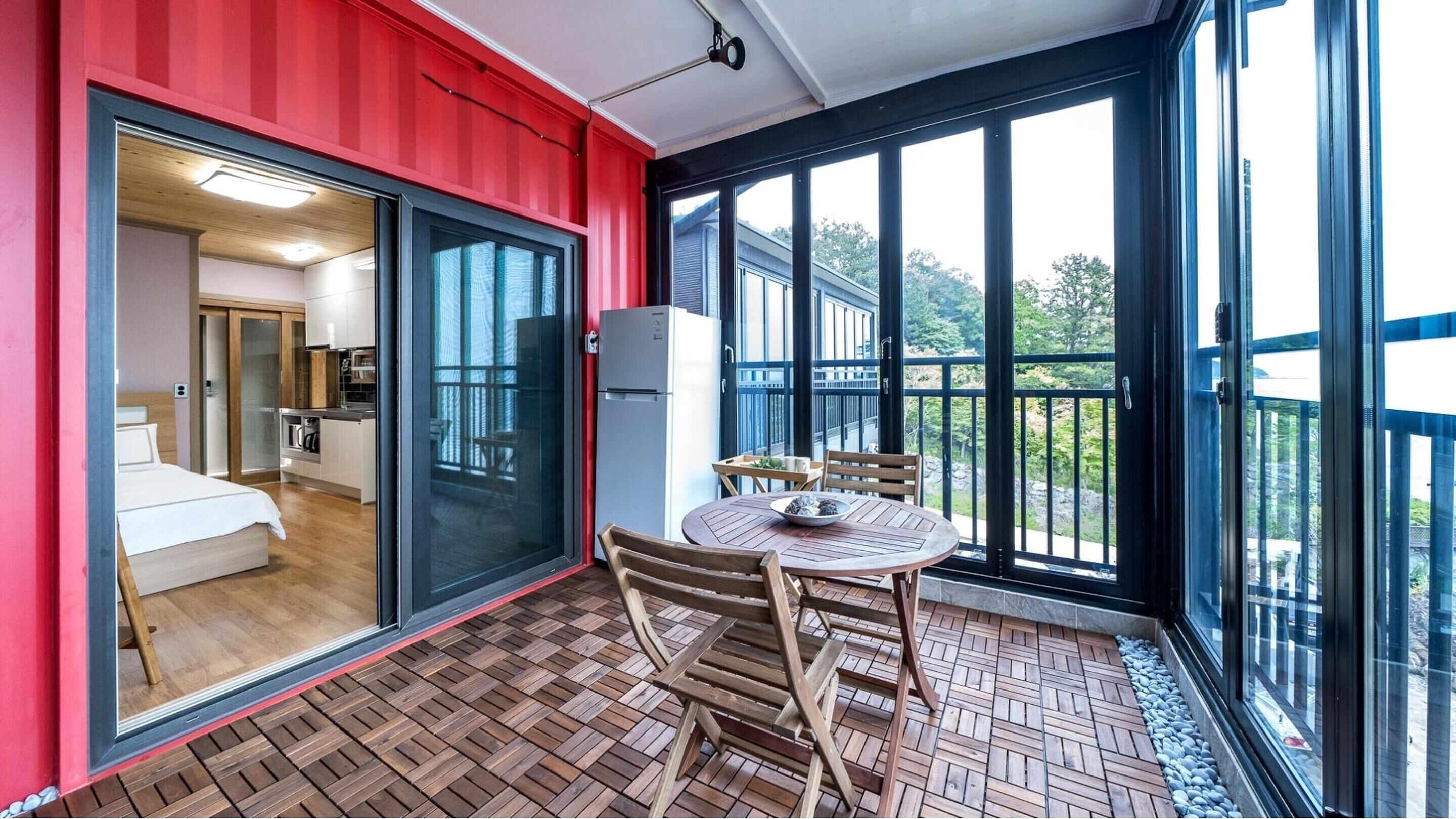 Una habitación moderna con puerta corrediza de vidrio que da a una cocina, un balcón con mesa y sillas, y un ventanal con vistas al exterior.