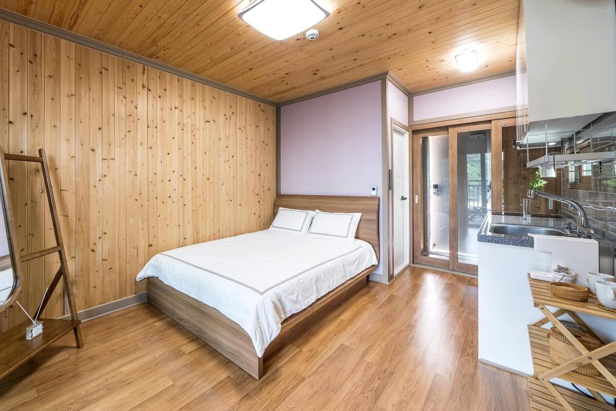 Une chambre avec un plancher en bois, un lit avec une literie blanche, une armoire en bois et une salle de bain dotée d’une cabine de douche en verre.