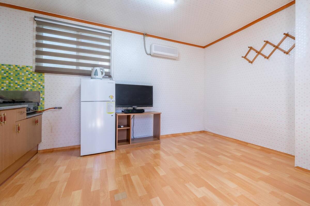Eine kleine Küche mit Kühlschrank, Herd und Mikrowelle. Ein Fenster mit Jalousien, ein Fernseher auf einem Standfuß und ein Holzboden.