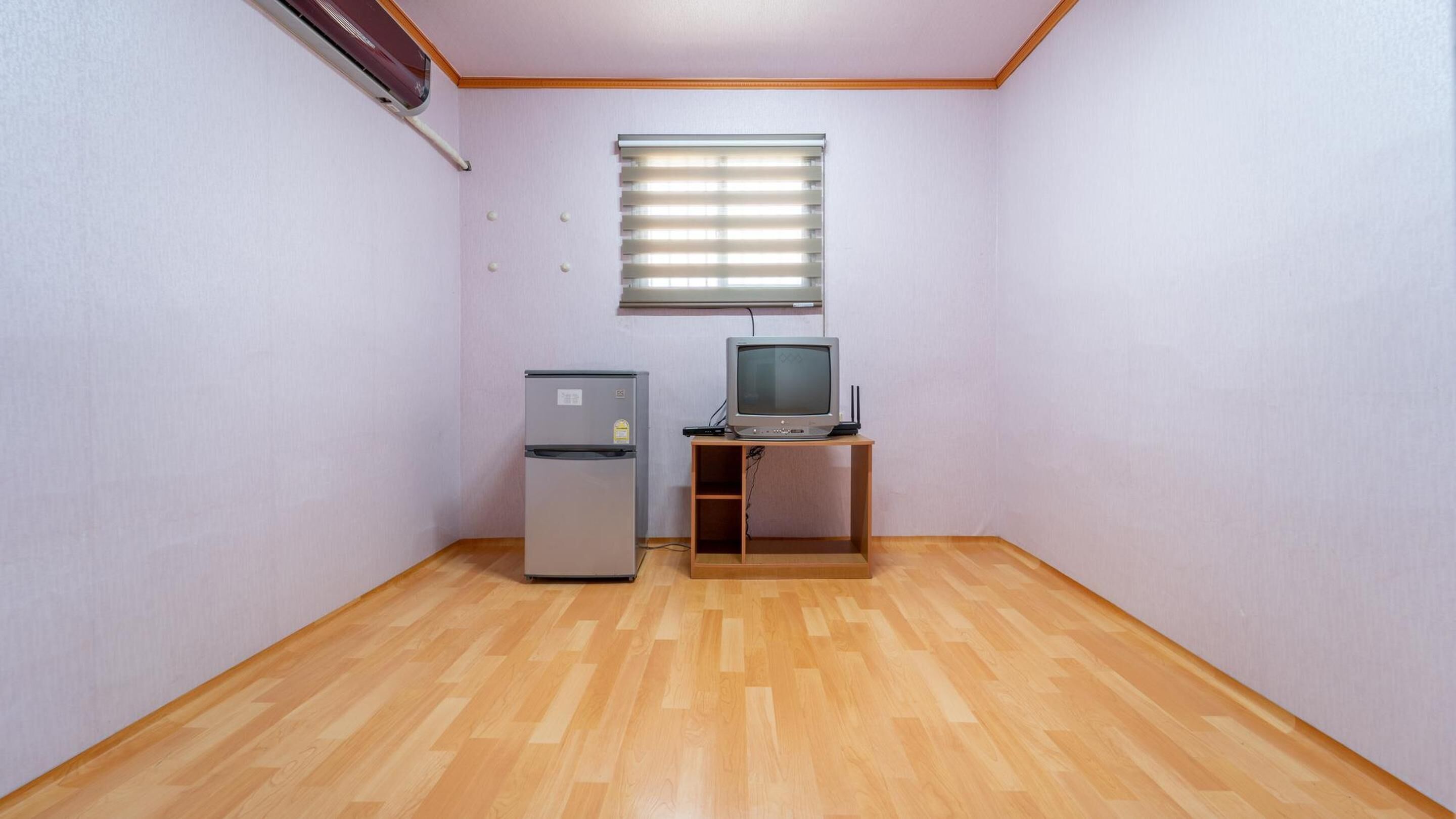 Una habitación pequeña con suelo de madera, un refrigerador, un televisor sobre un mueble de madera y una ventana con persianas.