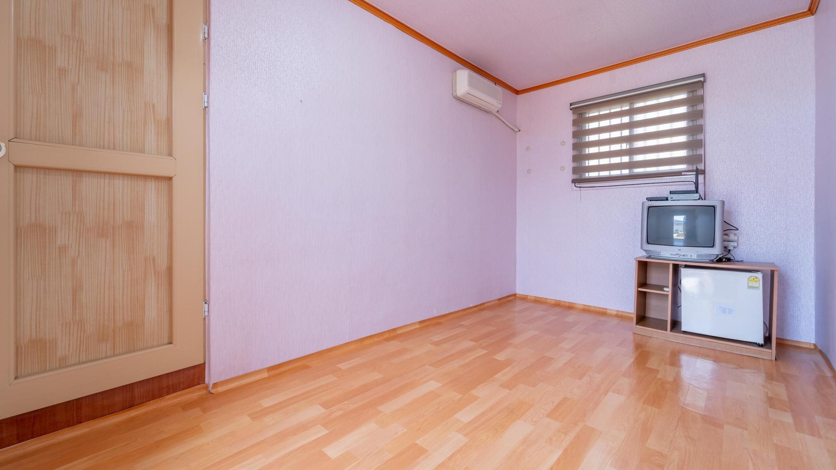 Quarto com piso de madeira, porta fechada, janela com persianas e televisão sobre um suporte.