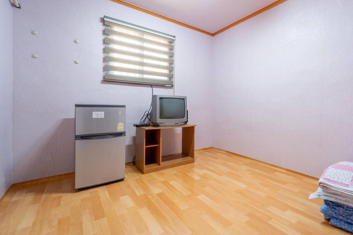 Ein kleines Zimmer mit Holzboden, einem Kühlschrank, einem Fernseher auf einem Holztisch und einem Bett mit einer Steppdecke.