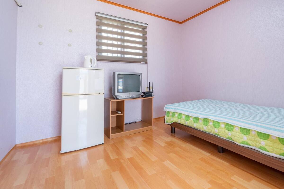 Ein kleines, ordentliches Zimmer mit einem Bett, einem Schreibtisch mit Computer und Kühlschrank sowie einem Fenster mit Jalousien.