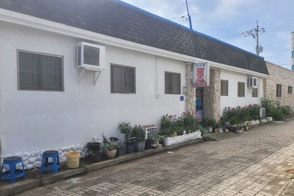 Un edificio blanco con varias ventanas, unidades de aire acondicionado y una puerta con un letrero. Hay plantas en macetas a lo largo de la acera y algunos taburetes.
