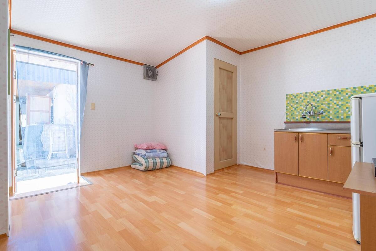 Ein kleiner, heller Raum mit Holzboden, einer Schiebetür zum Balkon, einer Küchenzeile mit gefliestem Spritzschutz und einer Tür zu einem weiteren Raum.