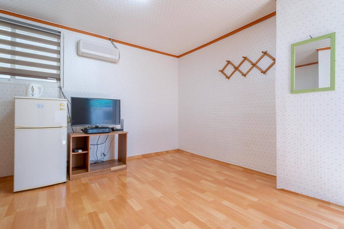 Ein kleiner, einfach eingerichteter Raum mit Holzboden, einem weißen Kühlschrank, einem Flachbildfernseher auf einem Holzständer und einer wandmontierten Klimaanlage.