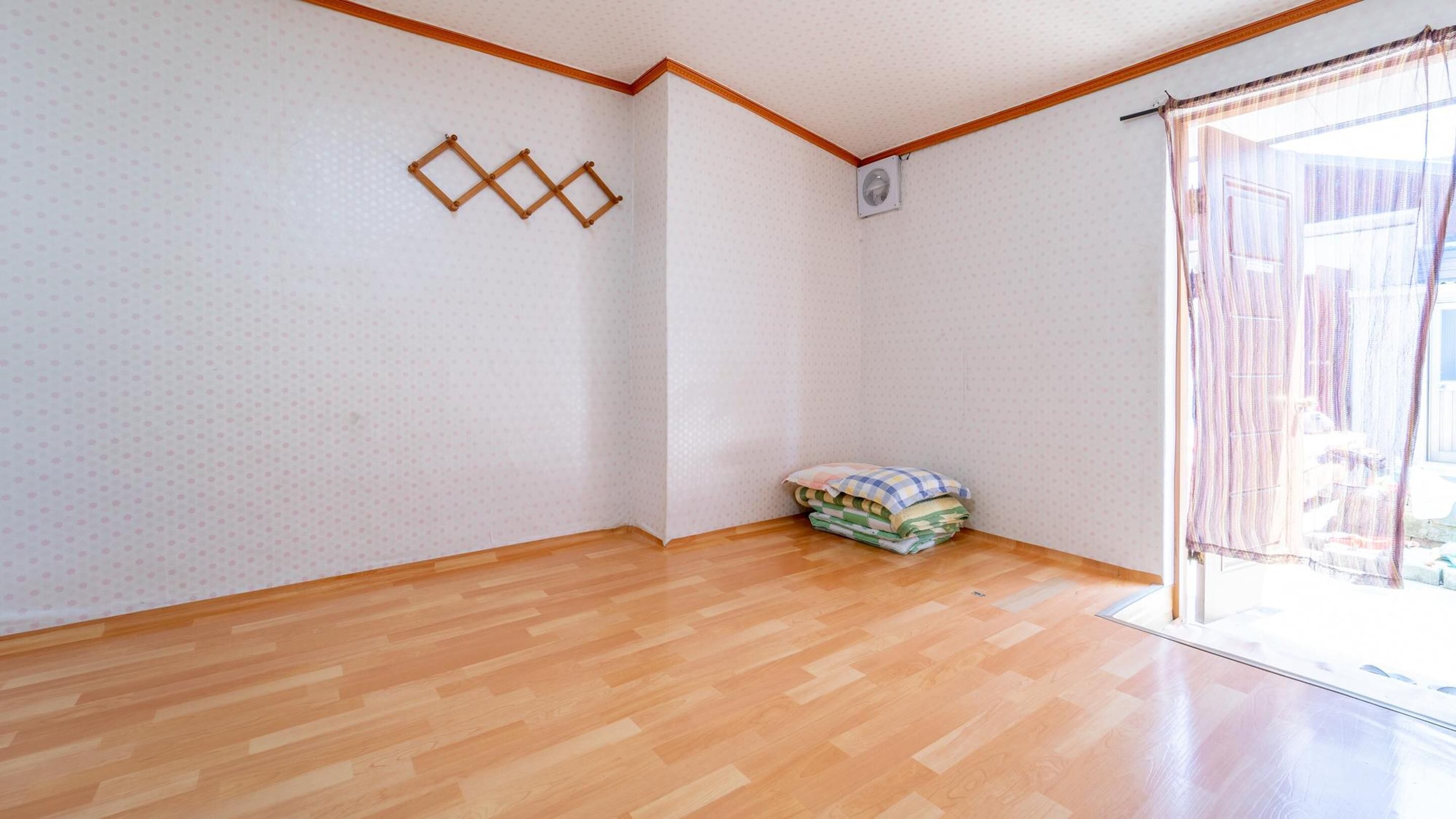 Una habitación con piso de madera, una pared angular con decoración geométrica, un montón de mantas dobladas y una ventana con cortinas.