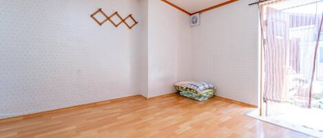Een kamer met houten vloer, een hoekwand met een geometrisch decor, een stapel opgevouwen dekens en een raam met gordijnen.