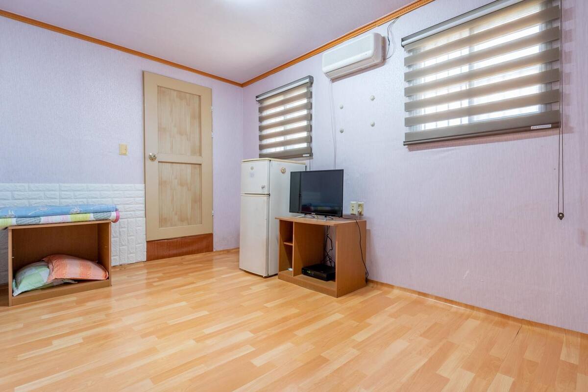 Ein kleiner, gut beleuchteter Raum mit Holzboden, einem Bett mit bunter Decke, einem Kühlschrank, einem Fernseher auf einem Holzschrank und zwei Wandklimaanlagen.